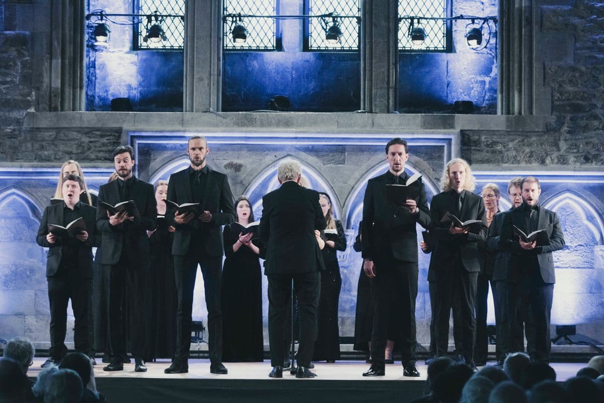 Conductor Mogens Dahl leads his chamber choir, standing with his back to the audience and conducting with precision and passion. The image captures Dahl in an active conducting role, clearly demonstrating his engagement and skill. The choir performs energetically under his guidance, highlighting his role as artistic director and his dedication to creating exceptional musical experiences. This visual representation of Dahl in action emphasizes his focus on quality and aesthetics in music.