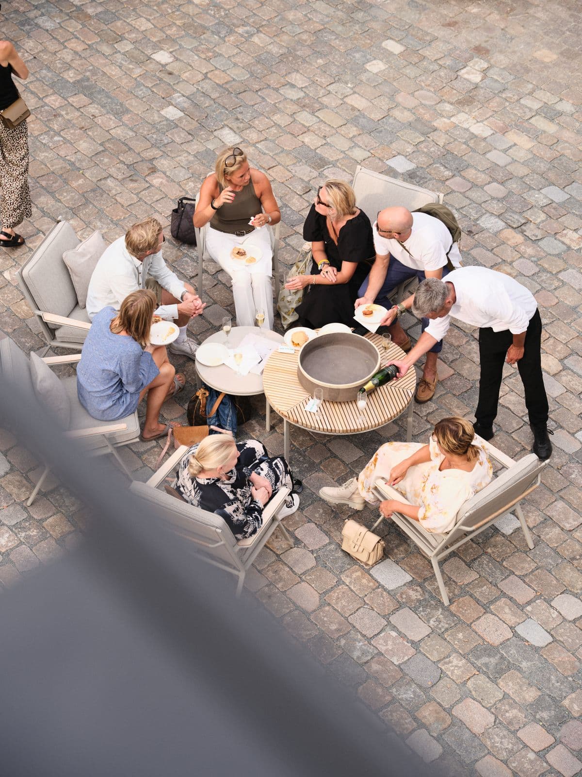 Image of guests sitting around a table outdoors, mingling, snacking, and drinking during an event at Mogens Dahl Concert Hall. The picture showcases a relaxed and social atmosphere outside.